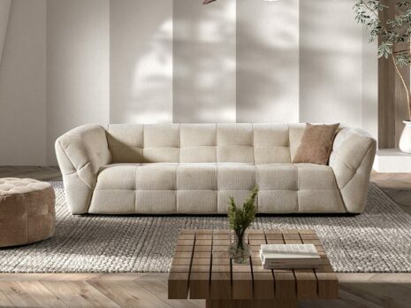 Canapé 2,5 places beige matelassé et rembourré dans un salon sur un tapis gris et devant un mur blanc avec une ombre d'une feuille de palmier