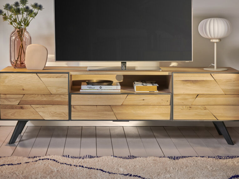 Meuble TV bas en bois massif à façade géométrique avec cadre noir, posé sur pieds inclinés, agrémenté de décoration, livres et lampe design dans un salon cosy.