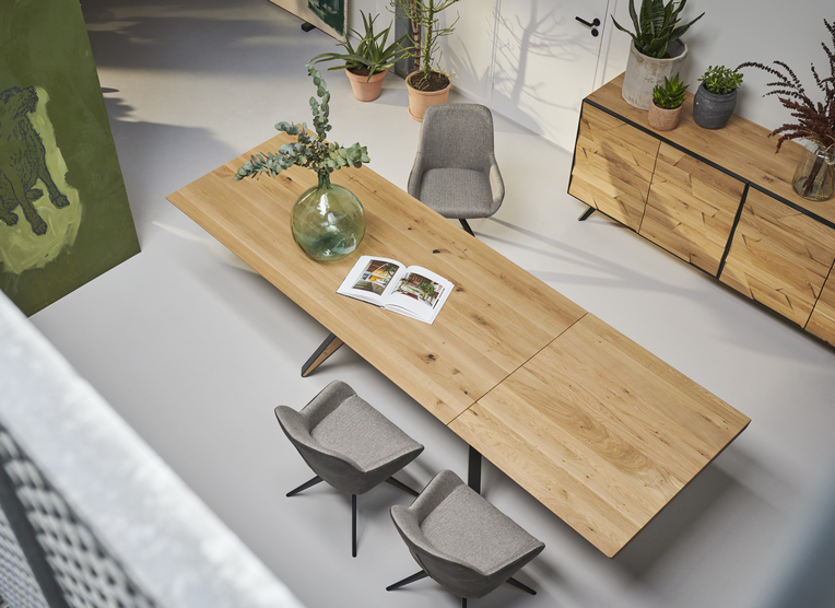 Grande table à manger rectangulaire en bois clair avec pieds métalliques noirs, décorée d’un vase en verre vert et d’un magazine ouvert. Trois chaises grises entourent la table, placée dans une salle moderne avec un buffet en bois et plusieurs plantes vertes en pots.