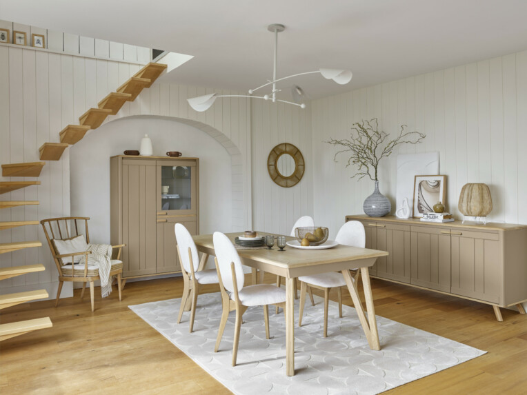 Salle à manger au style scandinave avec une table rectangulaire en bois clair, entourée de chaises blanches aux pieds en bois. La pièce est décorée avec un buffet assorti, un vaisselier, des objets décoratifs naturels et un grand tapis clair. Un escalier en bois flottant et un fauteuil en bois avec coussin complètent l’ensemble.